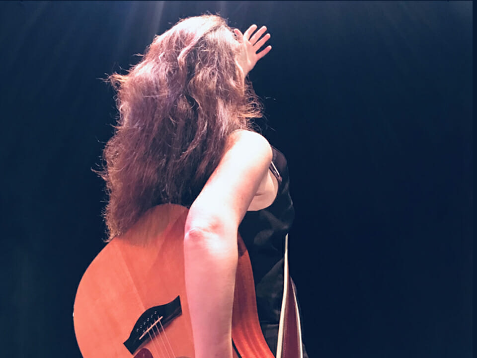 L'artiste Hélène Palardy se trouve de dos sur scène avec une guitare dans les mains.