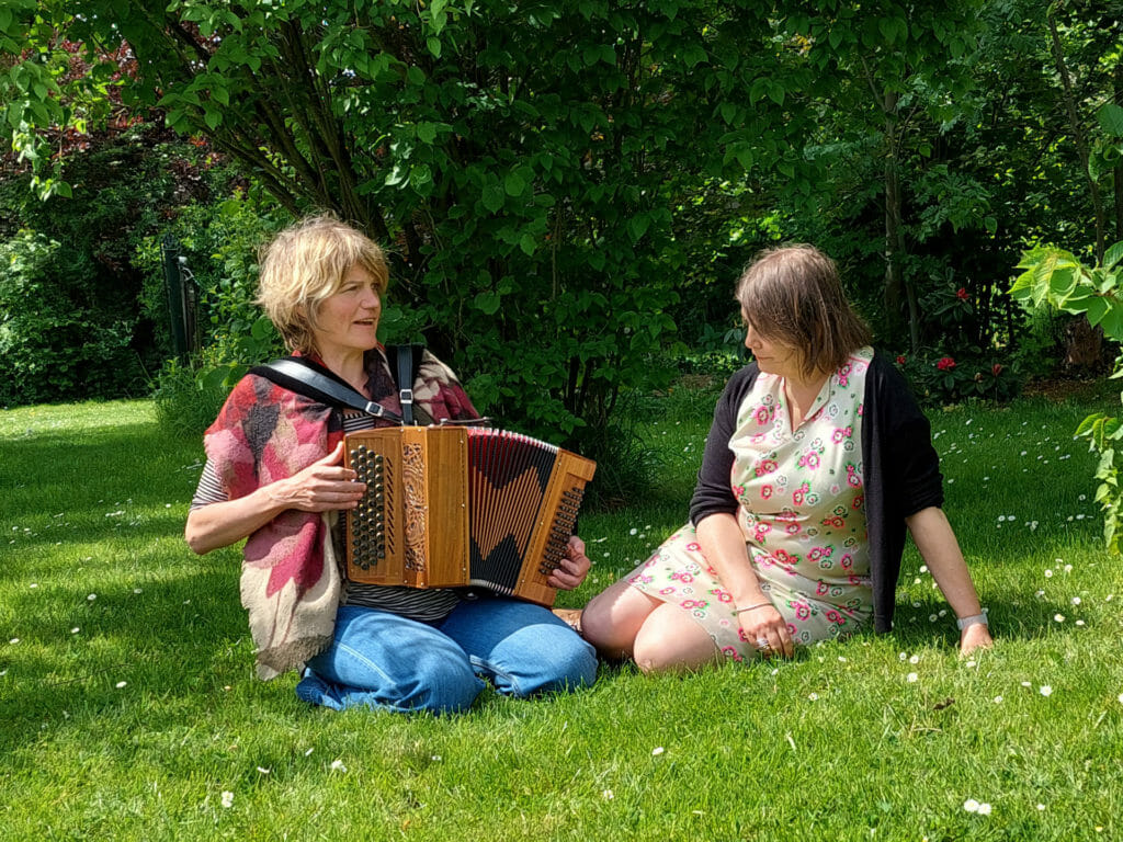 Deux dames sont assises sur l'herbe et chantent ensemble, l'une est accompagnée d'un accordéon.